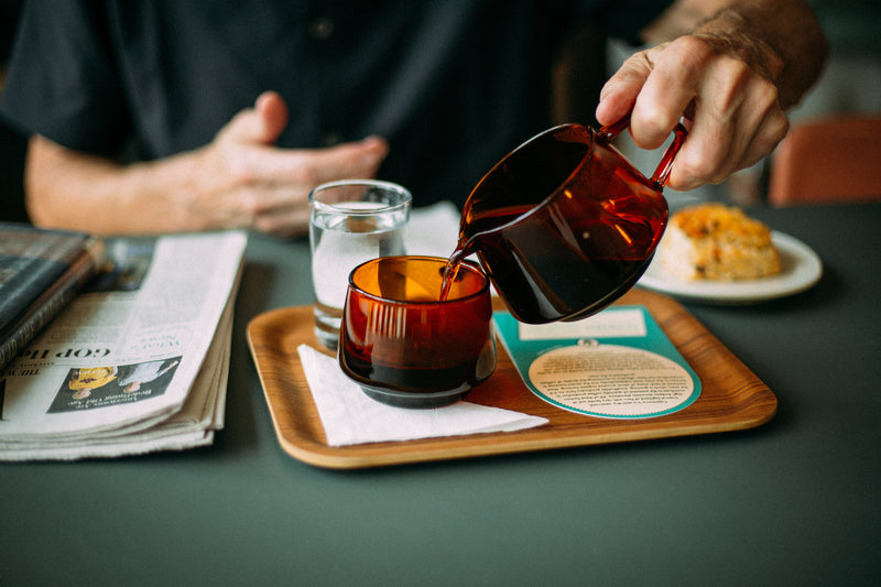Person pouring a dark liquid from an amber glass into another glass on a wooden tray with a newspaper and food items.